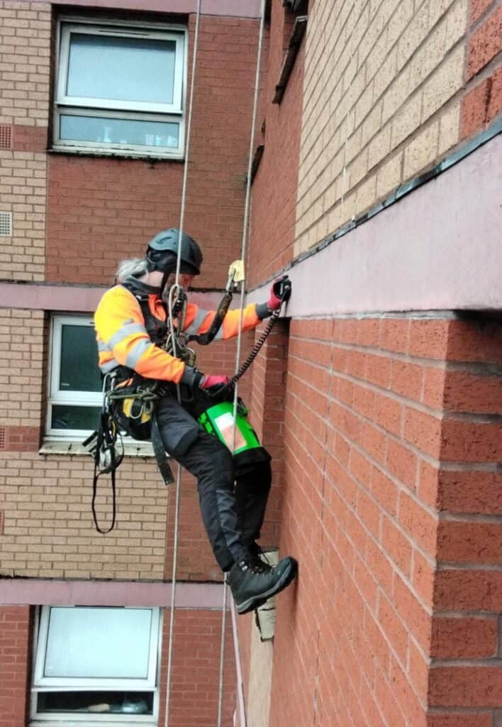 ORB_Rope_Access_Inspection2 | ORB Rope Access Rope-access technician in a bright orange vest and helmet rappelling down a brick building with safety gear and a harness.