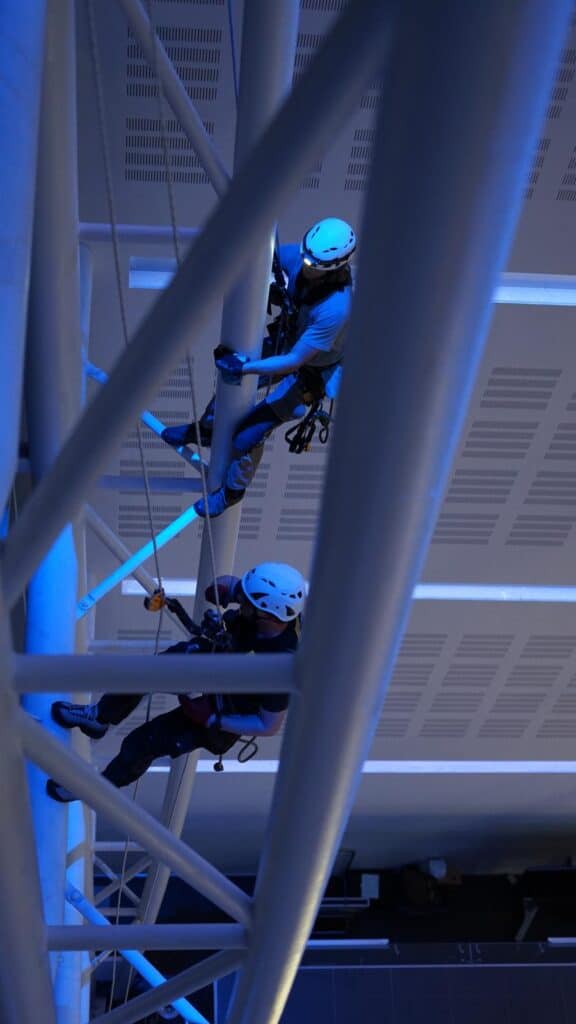 ORB_Rope_Access_Inspection3 | ORB Rope Access Two climbers in helmets and harnesses ascend a steel framework inside a building, using ropes and anchors for safety.